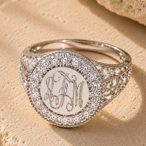 May include: A silver-toned ring featuring a circular face with an engraved monogram of the letters LJM. The center is surrounded by a halo of small clear stones, with additional stones set along the intricate, openwork filigree band.