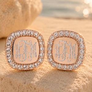 May include: A pair of rose gold square stud earrings with rounded corners, featuring a central engraved monogram with the letters JM and a border of small, clear, sparkling stones set into the metal frame of each individual earring piece.