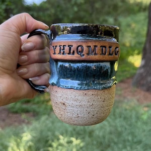 May include: Handmade ceramic mug with a black handle and rim, featuring a textured tan base and a dark teal and brown glazed body. The mug has the letters "YHLQM DLC" carved into the brown section. A unique, handcrafted piece for enjoying beverages.