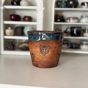 Könnte beinhalten: Eine handgefertigte Keramiktasse mit brauner, strukturierter Außenseite und einem dunkelblau-grünen glasierten Rand. Die Tasse zeigt geätzte Leopardenköpfe um den Körper. Die Tasse ist ein einzigartiges Keramikstück.