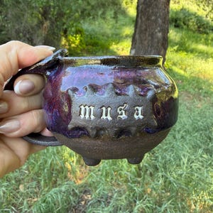 May include: A handcrafted ceramic mug with a dark brown base and a deep purple and maroon glaze. The word "musa" is inscribed in white letters on the front. The mug has a unique handle and three small feet.
