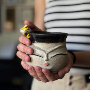May include: A handmade ceramic mug with a face design. The mug has a dark brown rim, a beige body, and a face with closed eyes and pink lips. A yellow and white bee is attached to the side. The mug is held in both hands.