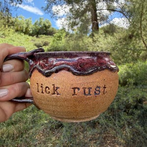May include: A handmade ceramic mug with a brown exterior and a dark red, glossy rim. The words "lick rust" are carved into the mug's surface. The mug has a unique handle and is set against a natural outdoor backdrop.