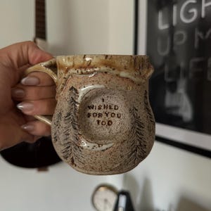 May include: A speckled ceramic mug with a textured surface and a unique handle. The mug features a forest design with trees and the text "I WISHED FOR YOU TOO". The mug is held by a person.