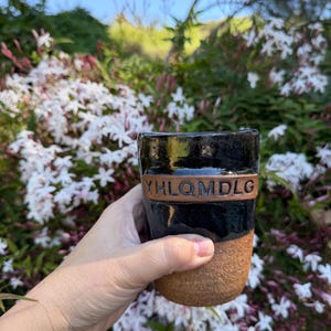 May include: A handmade ceramic mug with a black glazed top and a textured brown base. The mug features a brown band with the letters "YHLQMDLG" embossed on it. The mug is held in front of a backdrop of white flowers and greenery.