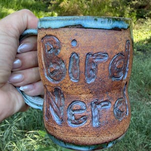 May include: A handmade ceramic mug with the words "Bird Nerd" carved into the brown surface. The rim and handle have a blue-green glaze. The mug is held against a backdrop of green foliage.