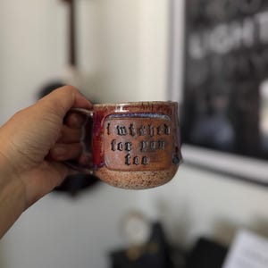 May include: A handmade ceramic mug with a speckled tan and deep red glaze. The mug features a rectangular embossed panel on the front with the text "i wished for you too" in a dark, gothic-style font. It has a curved handle on the left side.