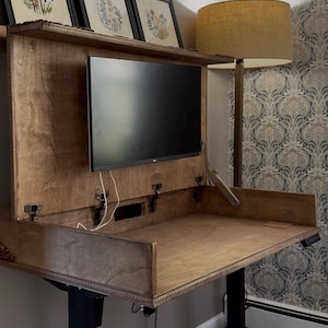 Op de afbeelding: Een houten sta-bureau met een ingebouwde monitorbeugel en een scharnierend achterpaneel. Het bureau heeft een donkere houten afwerking, zwarte metalen poten en een vlak werkoppervlak. Een zwarte computermonitor is in het midden bevestigd.