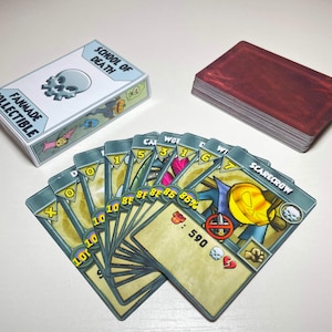 May include: A fanmade collectible card game titled School of Death featuring a deck box with a skull icon, a stack of red-backed cards, and a fanned-out hand of game cards displaying various character illustrations, numerical values, and icons.