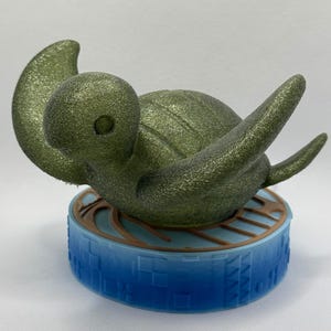 May include: A glittery olive green sea turtle figurine with a rounded shell and extended flippers, mounted on a circular base that features a gradient of blue with a textured grid pattern and a brown circular accent on the top surface.