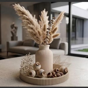 Peut inclure: Un vase cylindrique beige à côtes repose sur un plateau circulaire assorti également côtelé. Le vase contient de grandes herbes de la pampa plumeuses et des tiges séchées. Le plateau est orné de capsules de pavot, de petites pommes de pin.