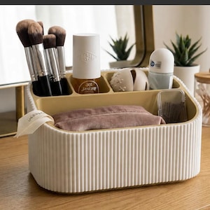 Peut inclure: Un organisateur de bureau ovale de couleur crème avec un extérieur nervuré vertical et un intérieur beige. Il comporte plusieurs compartiments contenant des pinceaux de maquillage, un flacon Sol de Janeiro, une petite sphère beige et une pochette en velours.