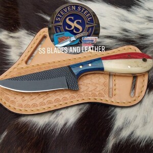 May include: A fixed-blade knife with a textured black metal blade and a handle featuring red, white, and blue segments. The knife rests on a tan leather sheath with decorative stamped patterns and stitched edges, featuring the text SS BLADES AND LEATHER.