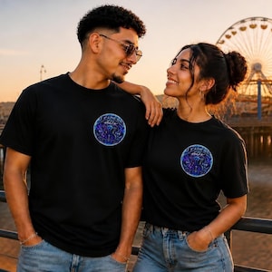 May include: A black short-sleeved t-shirt featuring a circular graphic on the left chest. The design contains blue and purple celestial patterns with the text "IT FADES TO DARK" printed in white lettering across the centre of the circular emblem.