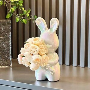 May include: A ceramic rabbit figurine with an iridescent, pearlescent finish. The rabbit is standing upright and holding a large bouquet of pale pink and cream-colored artificial peony flowers in its arms, featuring long ears and a smooth surface.