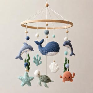 Könnte beinhalten: Ein hängendes Mobile mit Filz-Meeresbewohnern, darunter ein blauer Wal, zwei Delfine, eine Meeresschildkröte, ein Seestern, eine Krabbe und eine weiße Muschel. Die Figuren hängen an weißen Schnüren mit Filzkugeln an einem runden Holzrahmen.