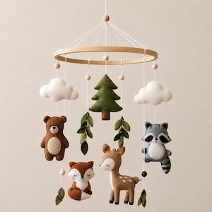 Può includere: Una giostrina per culla con una cornice circolare in legno e fili bianchi che sospendono figure in feltro di un orso bruno, una volpe, un cerbiatto, un procione, un pino verde, nuvole bianche e foglie verdi, con piccole perline di legno.