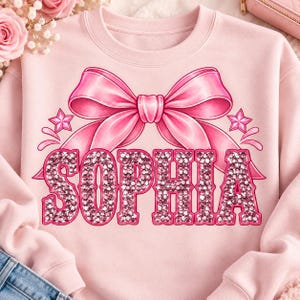 Peut inclure: Un sweat-shirt à col rond rose clair arborant un grand motif de nœud rose avec deux petites étoiles de chaque côté. Sous le nœud, le nom SOPHIA est imprimé en grandes lettres grasses avec un motif texturé rappelant des strass roses.