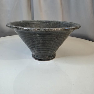 May include: A wide, conical ceramic bowl with a dark gray, textured glaze. The bowl features a prominent, flat, outward-curving rim and a narrow, circular base. The exterior surface displays subtle horizontal ridges and a mottled, uneven finish.