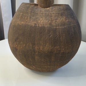May include: A round, dark brown ceramic vase with a textured, matte finish. The surface features etched, concentric wavy lines forming an organic shape in the center, with horizontal lines below and vertical ribbing on the narrow neck.