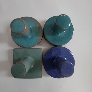 May include: Four ceramic stamps with cylindrical handles and flat bases. Three stamps have circular bases in teal, dark teal, and deep blue with a crackle glaze. One stamp has a square base in a muted sage green color. All feature smooth, glossy finishes.