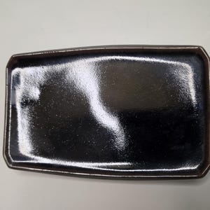 May include: A rectangular ceramic tray with a glossy, deep black finish and a dark brown rim. The surface features a smooth, reflective glaze with subtle variations in texture, creating a polished appearance with defined, slightly angled edges.