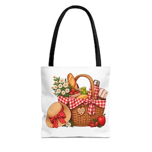 May include: A white tote bag with black handles featuring a graphic of a woven picnic basket filled with a baguette, strawberries, grapes, daisies, and a red checkered thermos. A straw hat with a red polka dot ribbon sits next to the basket.