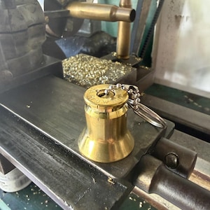 May include: A brass keychain featuring a flared base and a top section fashioned from a spent cartridge casing. The top of the casing is stamped with the text '30-06 SPRG' and 'Browning', and it includes a silver-toned metal ball chain and ring.