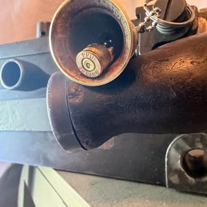 May include: A brass bell with a metallic clapper made from a PPU 30 CARB cartridge casing. The bell is attached to a silver-colored ball chain and a split ring. The bell is positioned in front of a dark brown wooden handle and a black rectangular object.