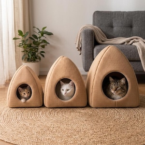 May include: Three tan, waffle-textured, triangular pet caves of varying sizes arranged in a row. Each cave features a circular opening. A small calico kitten occupies the smallest, a white cat sits in the middle, and a tabby cat rests in the largest.