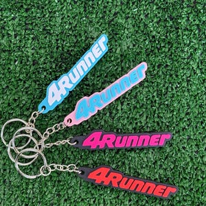 May include: Four flexible keychains featuring the text 4Runner in raised lettering. The set includes a light blue, a pink, a black with magenta text, and a black with red text keychain, each attached to a silver metal split ring and chain.