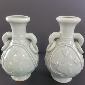 Puede incluir: Un par de jarrones de celadón verde pálido a juego con cuerpo bulboso, cuello acampanado y dos asas laterales con anillos adjuntos. La superficie exterior está decorada con un patrón en relieve de motivos florales y hojas.