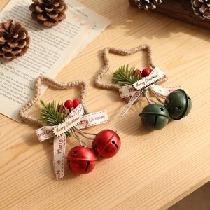May include: Two star-shaped ornaments made of twisted jute rope, each decorated with a small green pine sprig, red berries, and a white ribbon printed with the text Merry Christmas. One ornament features two red jingle bells, the other two green bells.