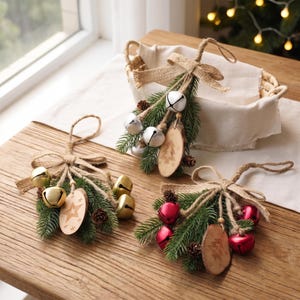 May include: Three decorative hanging ornaments featuring bundles of green pine needles, small brown pinecones, and jute twine bows. Each ornament includes metallic jingle bells and a small wooden disc engraved with either a star or a tree design.