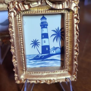 May include: A small rectangular picture frame with a metallic gold finish and a decorative bow at the top. The frame contains a blue and white illustration of a lighthouse, a small house, and palm trees situated on a coastal shoreline.