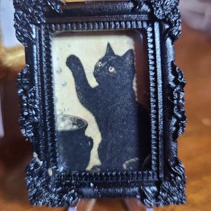 May include: A miniature rectangular black frame with ornate, textured edges containing an illustration of a black cat with yellow eyes reaching toward floating bubbles above a small black cauldron against a pale yellow background.