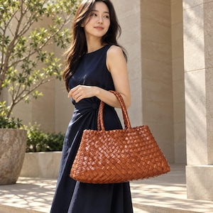 Puede incluir: Un bolso tote grande y rectangular confeccionado con tiras de cuero marrón entrelazadas. El bolso presenta una base estructurada, una abertura amplia y dos asas de cuero trenzado resistentes. El material tiene un acabado brillante, mostrando un patrón de tejido de cesta consistente en toda la superficie exterior del bolso.