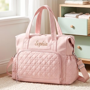 May include: A light pink diaper bag featuring a quilted front pocket, two top handles, and a detachable shoulder strap. The name Sophia is embroidered in a cursive font on the upper front panel. The bag includes side pockets and metal hardware.
