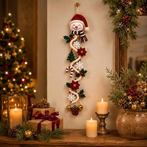 May include: A vertical crochet wall hanging featuring a snowman head with a red hat and striped scarf, connected by a cream-colored rope adorned with white snowflakes, red poinsettias, a green tree, a candy cane, and two gold bells.