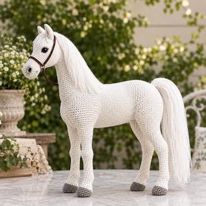 Pode incluir: Um brinquedo de cavalo feito em crochê com fio branco off-white, apresentando uma crina e cauda longas e fluidas. O cavalo possui uma rédea de fio castanho, olhos de segurança escuros e cascos cinzentos, numa pose clássica de perfil.