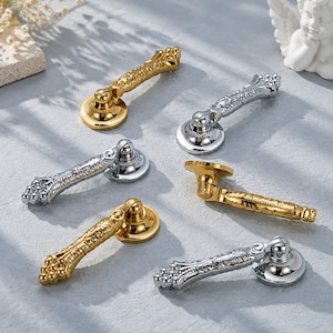 Puede incluir: Un conjunto de seis tiradores de cajón de metal con diseño de asa abatible y patrones florales en relieve. Tres tiradores tienen un acabado en tono dorado pulido y tres tienen un acabado en tono plateado pulido, cada uno con una base circular.