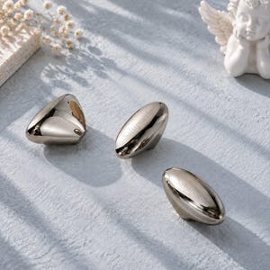 May include: Three polished metallic cabinet knobs with a smooth, reflective finish. Two knobs are elongated and oval-shaped, while one is triangular. Each piece features the brand name Salsipuede engraved on the surface in a subtle font.