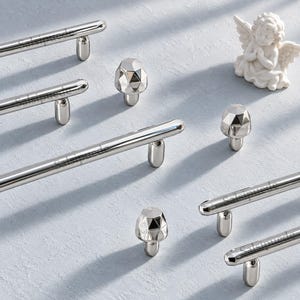 May include: A collection of polished silver-toned metal cabinet hardware, including several long, cylindrical bar pulls with rounded ends and three faceted, diamond-shaped knobs, arranged alongside a small white figurine of a winged angel.
