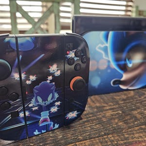 May include: A video game console controller and dock featuring a Sonic the Hedgehog graphic. The controller has black buttons, a black joystick with an orange ring, and a blue character design. The dock displays a large blue Sonic character face.