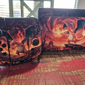 May include: A video game console dock and a pair of controllers featuring a graphic print of the orange dragon-like creature Charizard breathing fire against a dark, fiery landscape. The controllers include black joysticks and buttons.
