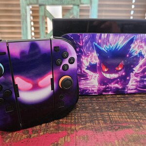May include: A video game console dock and controller set featuring a purple and black Gengar character skin. The controller has black buttons, a light blue left joystick, and an orange right joystick. The dock displays a glowing purple Gengar graphic.