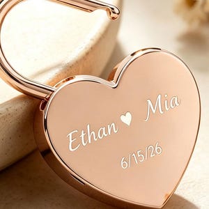 May include: A rose gold heart-shaped padlock with a polished metallic finish. The front surface is engraved with the names Ethan and Mia separated by a small heart symbol, followed by the date 15/06/26 in a clean, white, sans-serif font.