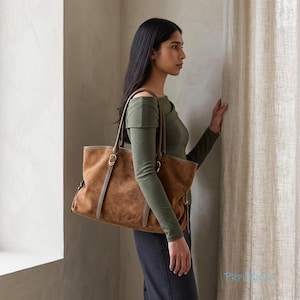 Puede incluir: Un bolso tote grande de ante marrón con dos correas para el hombro, herrajes de metal en tono dorado y detalles de cuero verticales. El bolso tiene un cuerpo rectangular espacioso con costuras laterales visibles y una base estructurada.