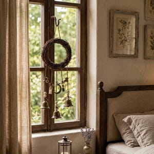 May include: A hanging wall decoration featuring a circular twig wreath adorned with dangling elements including small metal bells, glass vials, wooden beads, and a crescent moon charm, suspended by twine from a dark metal hook on a window frame.