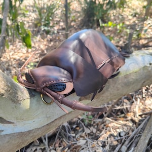 May include: A handcrafted leather sculpture shaped like a large insect, featuring a dark brown segmented body, visible stitching along the edges, black leather eye details, and two long antennae extending from the front with a small gold metal ring.